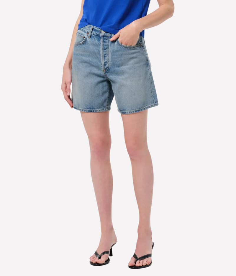 A model wearing light-wash blue denim shorts with a blue top and black heeled sandals, Agolde Dame Short in Reset