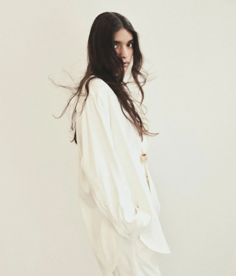 A woman with long, dark wavy hair wearing an oversized white long-sleeve top and matching white bottoms, photographed against a plain off-white studio background.