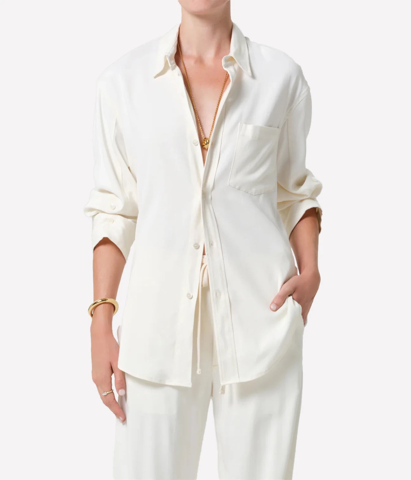 A model wearing a cream-colored linen button-up shirt and matching trousers, styled with gold jewelry against a plain white background.