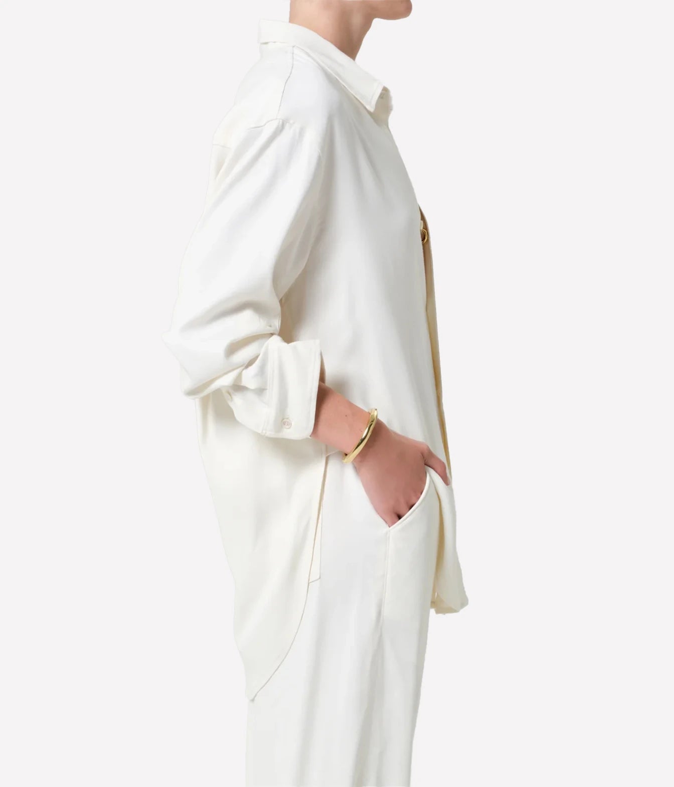 A side-profile view of a model wearing a long-sleeve, oversized off-white shirt jacket with gold-toned buttons, set against a plain light gray background.