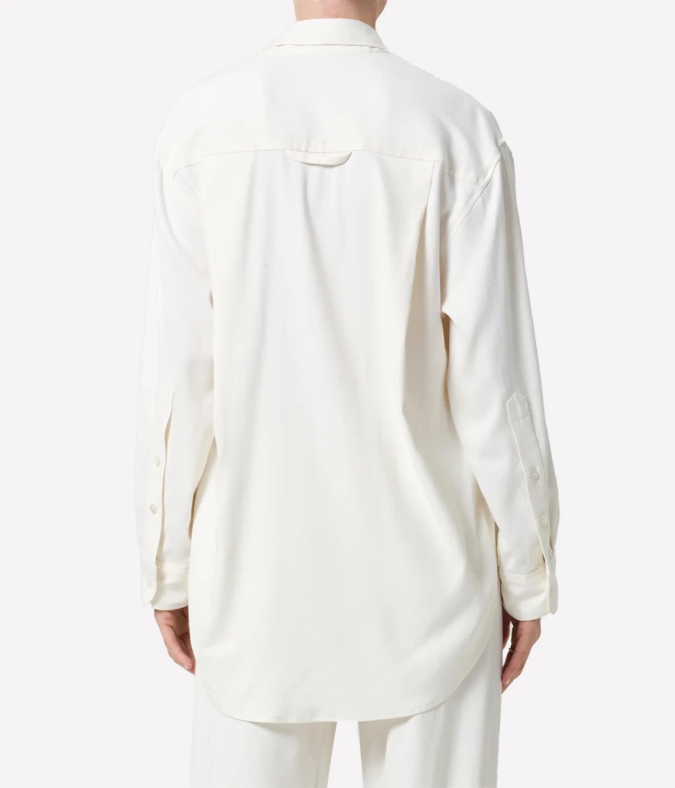A person shown from the back wearing a long-sleeve cream-colored button-up shirt and matching light pants against a plain light gray studio background.