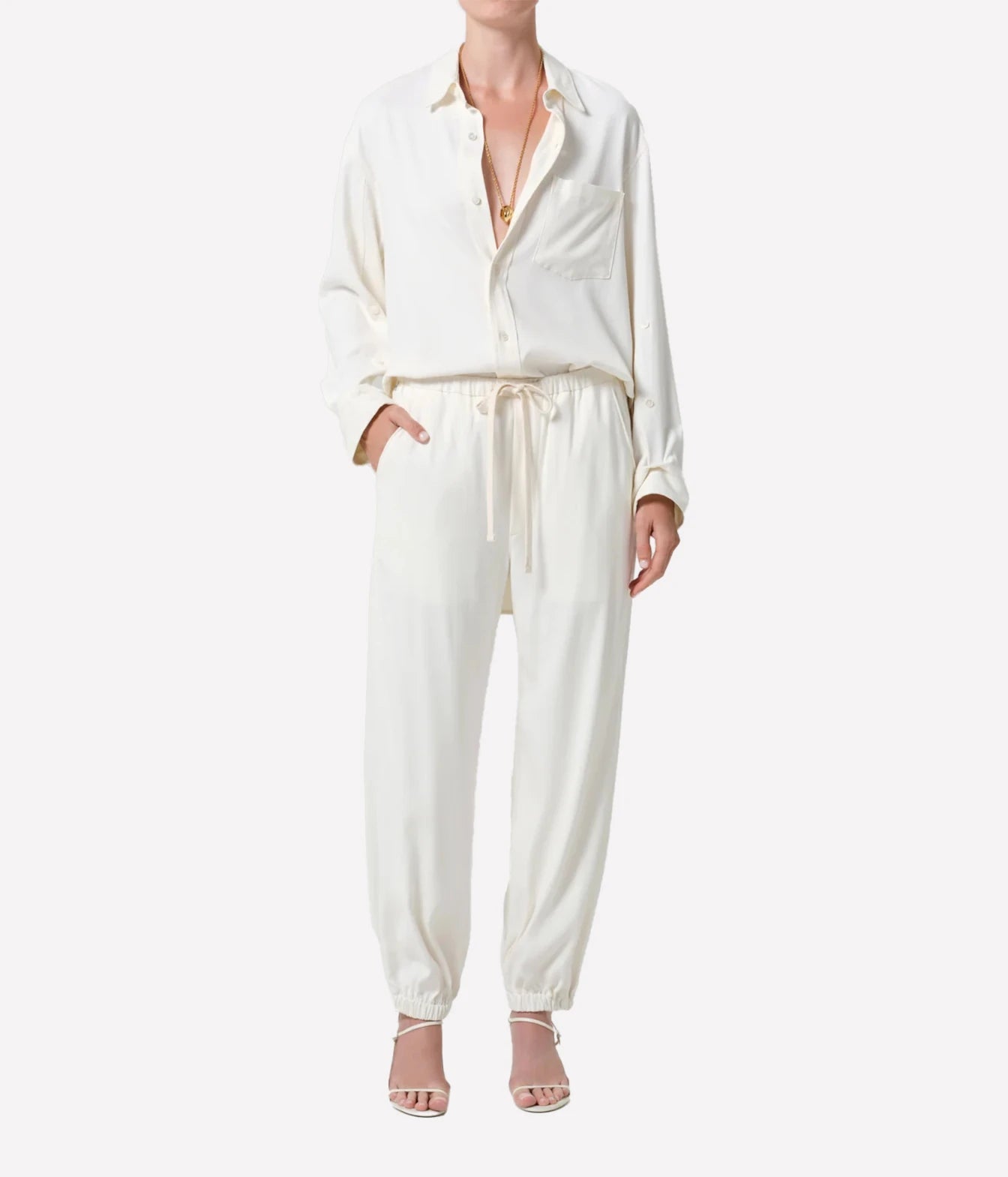A model wearing a cream-colored two-piece set consisting of a long-sleeve button-up shirt and matching drawstring trousers, paired with nude high-heeled sandals against a plain white background.
