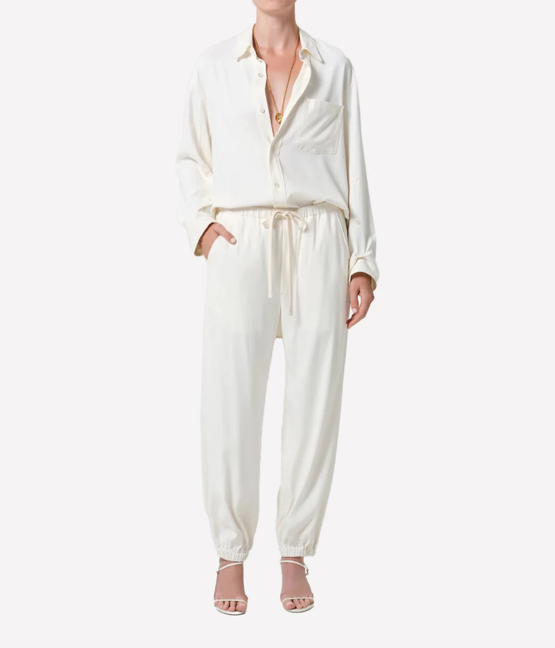 A model wearing a cream-colored two-piece set consisting of a long-sleeve button-up shirt and matching drawstring trousers, paired with nude high-heeled sandals against a plain white background.