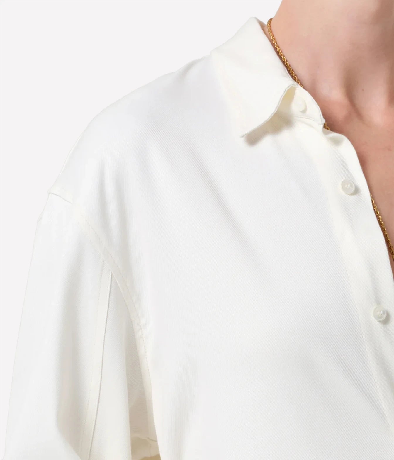Close-up of a person wearing a white button-up shirt with a pointed collar and white buttons, paired with a thin gold chain necklace, against a plain light background.