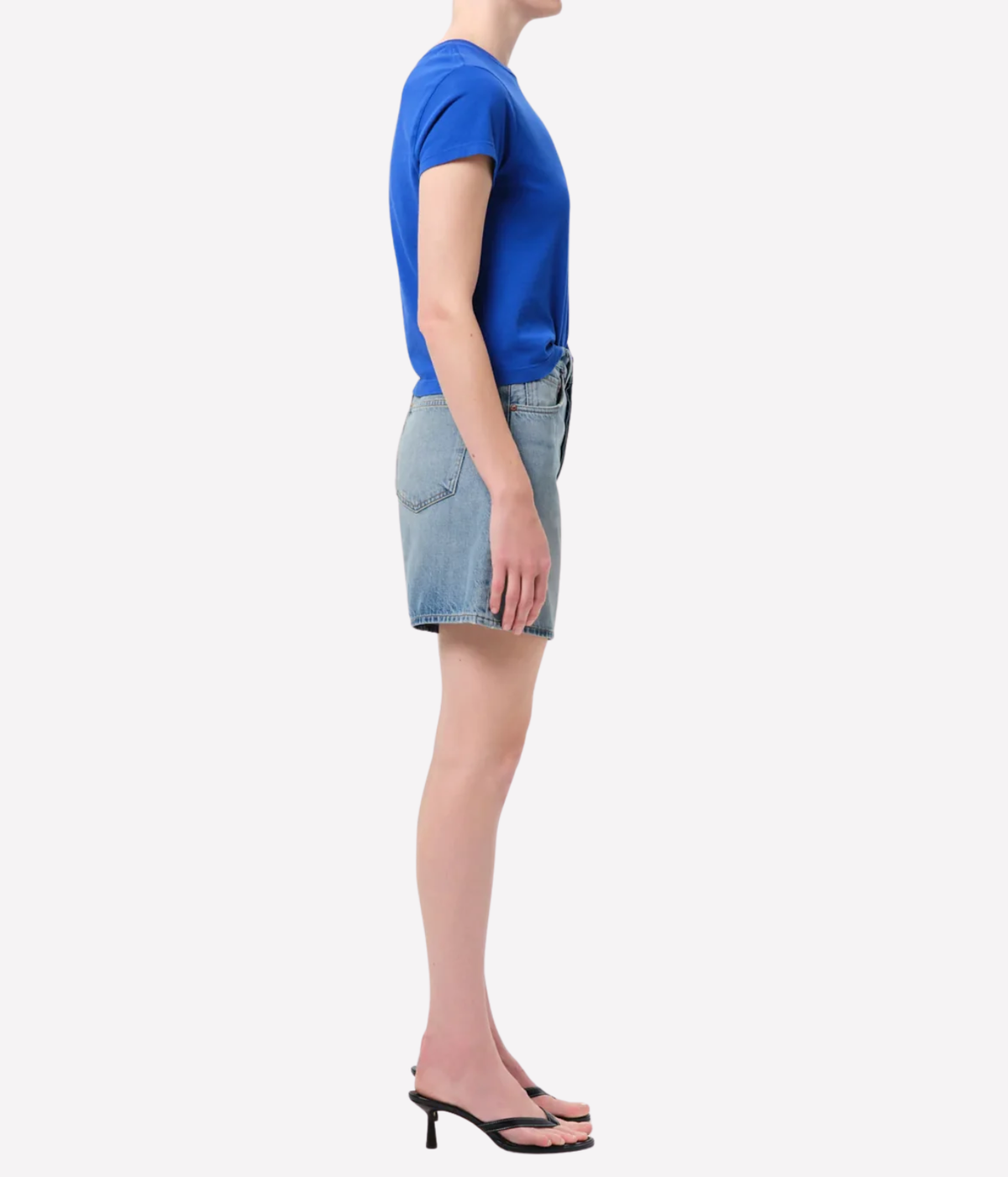 A side-profile view of a model wearing a bright blue short-sleeve t-shirt, light-wash denim shorts, and black slide sandals, Agolde Dame Short in Reset
