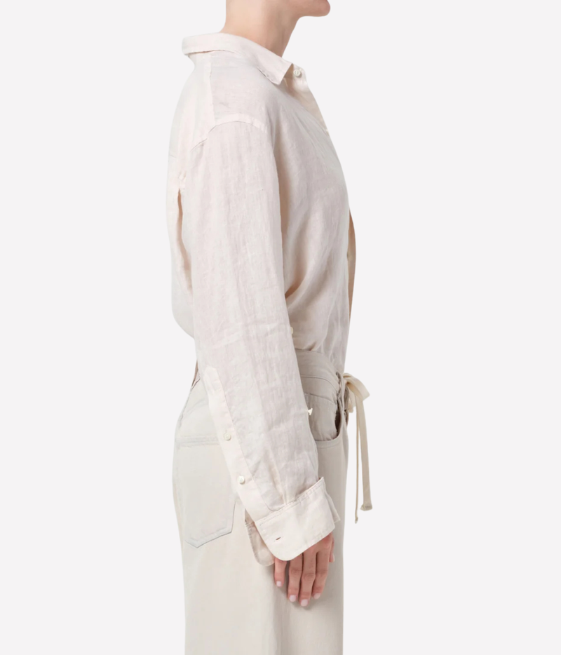 A side-profile view of a model wearing a light beige linen shirt with a collar, rolled cuffs, and a matching waist tie, paired with light beige trousers against a plain white background.
