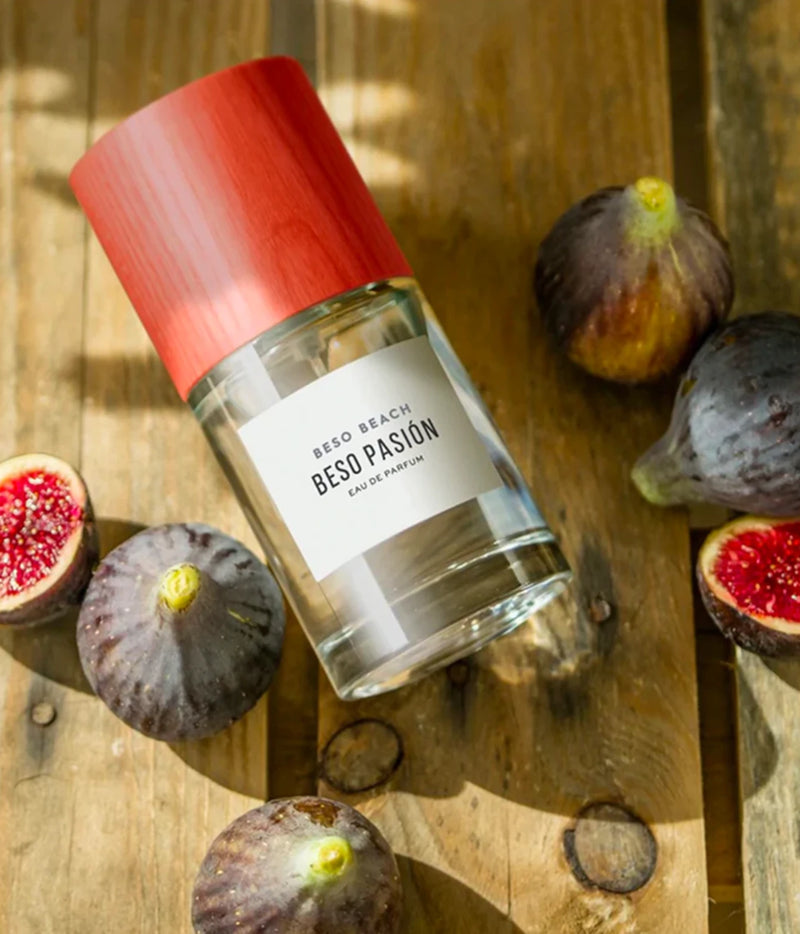 A clear glass perfume bottle with a red cap and a white label reading 'BEAD BEACH BESO PASIÓN' lies on a rustic wooden surface surrounded by whole and halved figs.