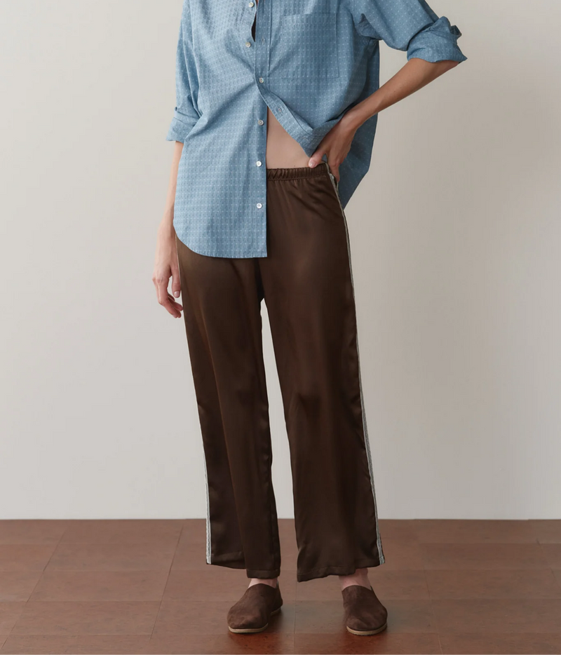 Front view of a model wearing the DONNI. The Silk Lace Stripe Crop Pant in Espresso with a blue printed button-up shirt worn half-open with rolled sleeves and brown suede flat slippers
