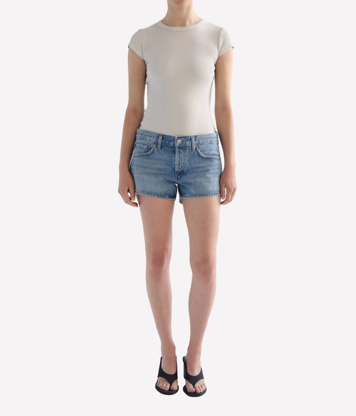 A model wearing a beige ribbed short-sleeve top and light-wash blue denim shorts, Agolde Low Rise Parker Short in Replica