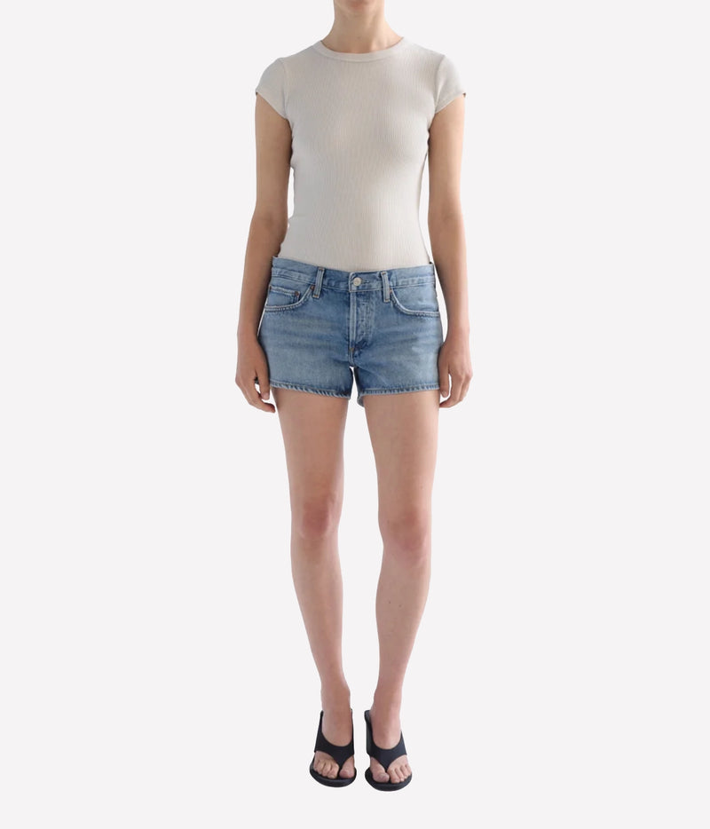 A model wearing a beige ribbed short-sleeve top and light-wash blue denim shorts, Agolde Low Rise Parker Short in Replica