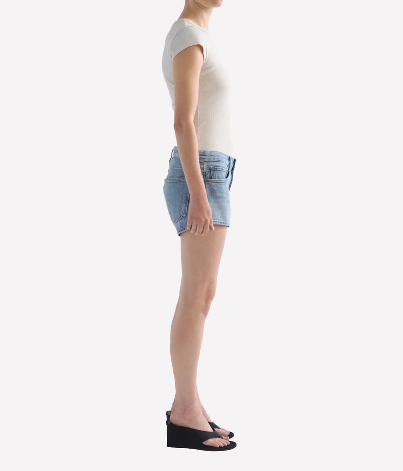 A side-profile view of a model wearing light-wash denim shorts, Agolde Low Rise Parker Short in Replica, a white short-sleeve top, and black wedge sandals