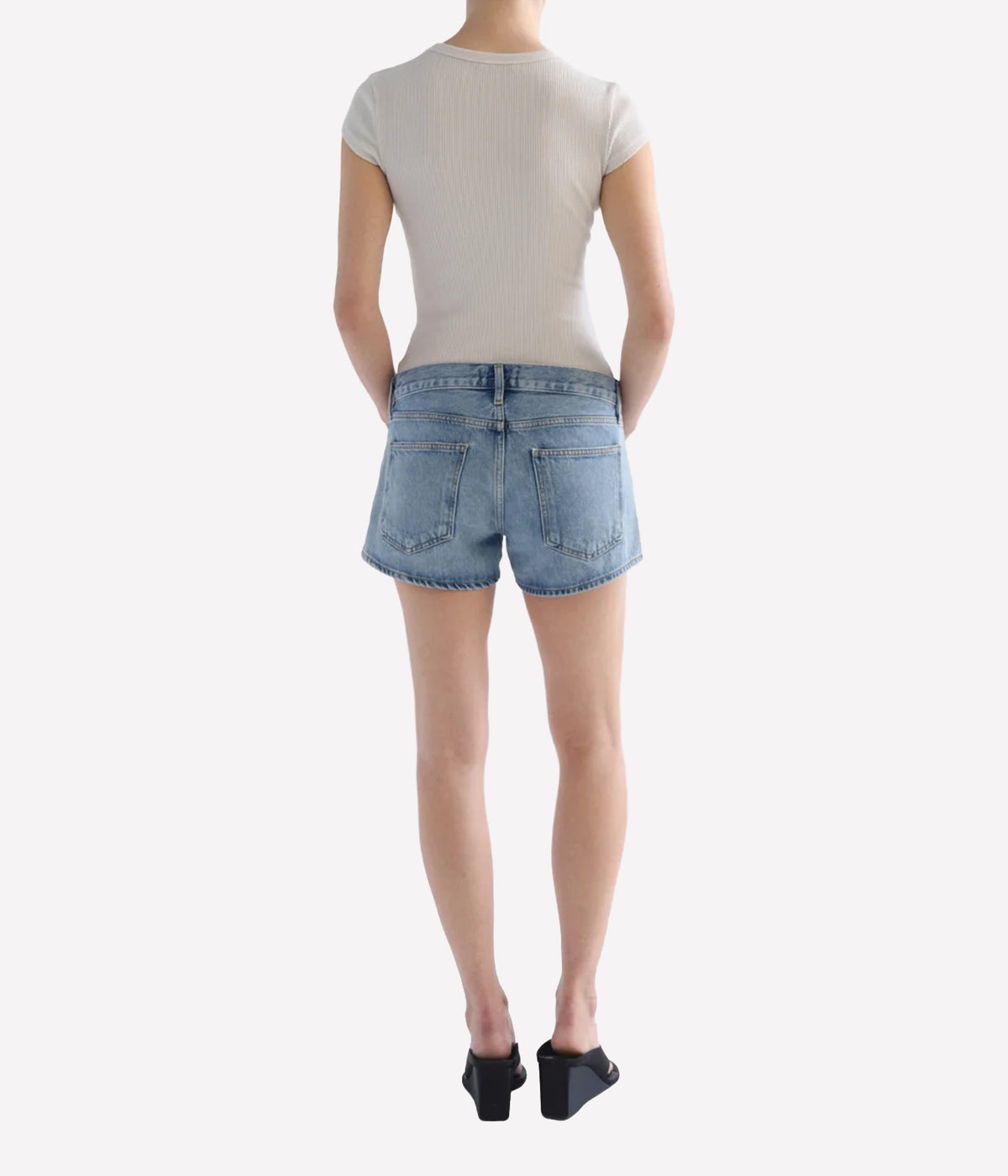 Rear view of a model wearing light-wash blue denim shorts, Agolde Low Rise Parker Short in Replica, a light beige ribbed short-sleeve top, and black wedge sandals