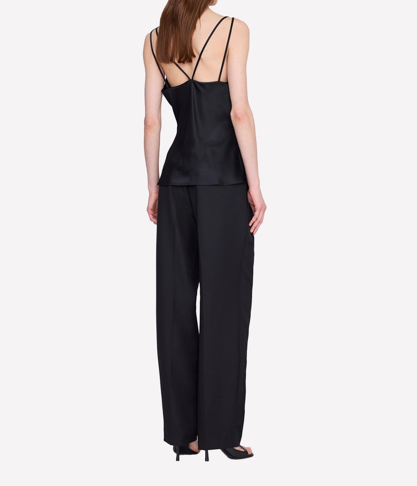 A model shown from the back wearing a black satin camisole top with thin straps and matching wide-leg trousers, paired with black high heels, against a light gray background.