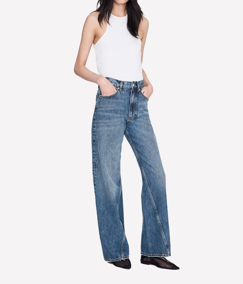 A model wearing a pair of high-waisted, wide-leg blue denim jeans with a white sleeveless top and black pointed shoes, shown against a plain light gray background.
