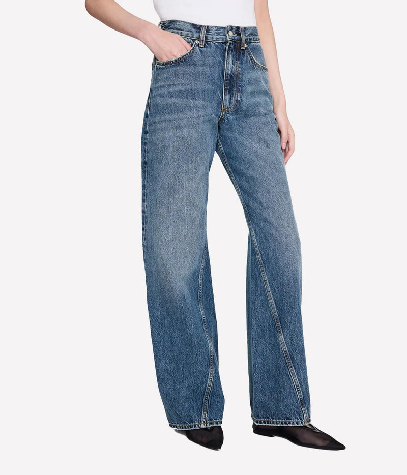 A model wearing a pair of high-waisted, wide-leg blue denim jeans with a faded wash, paired with a white top and black pointed flats, shown against a light gray studio background.