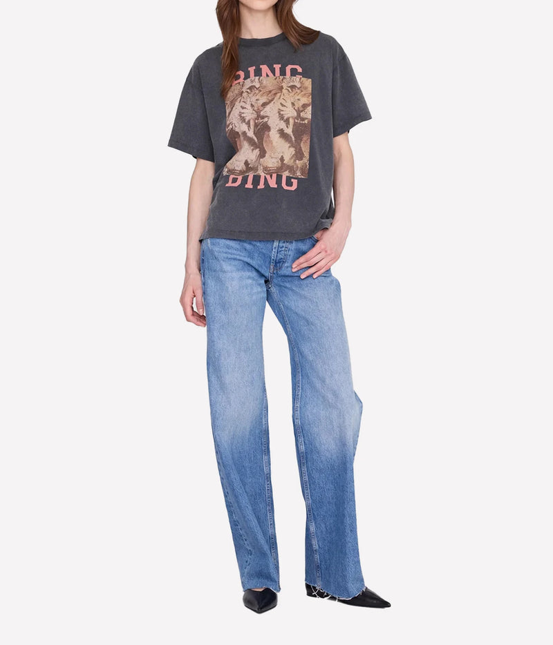 A model wearing a dark gray graphic t-shirt with a pink and beige wolf print, paired with wide-leg blue jeans and black pointed shoes, against a light gray background.