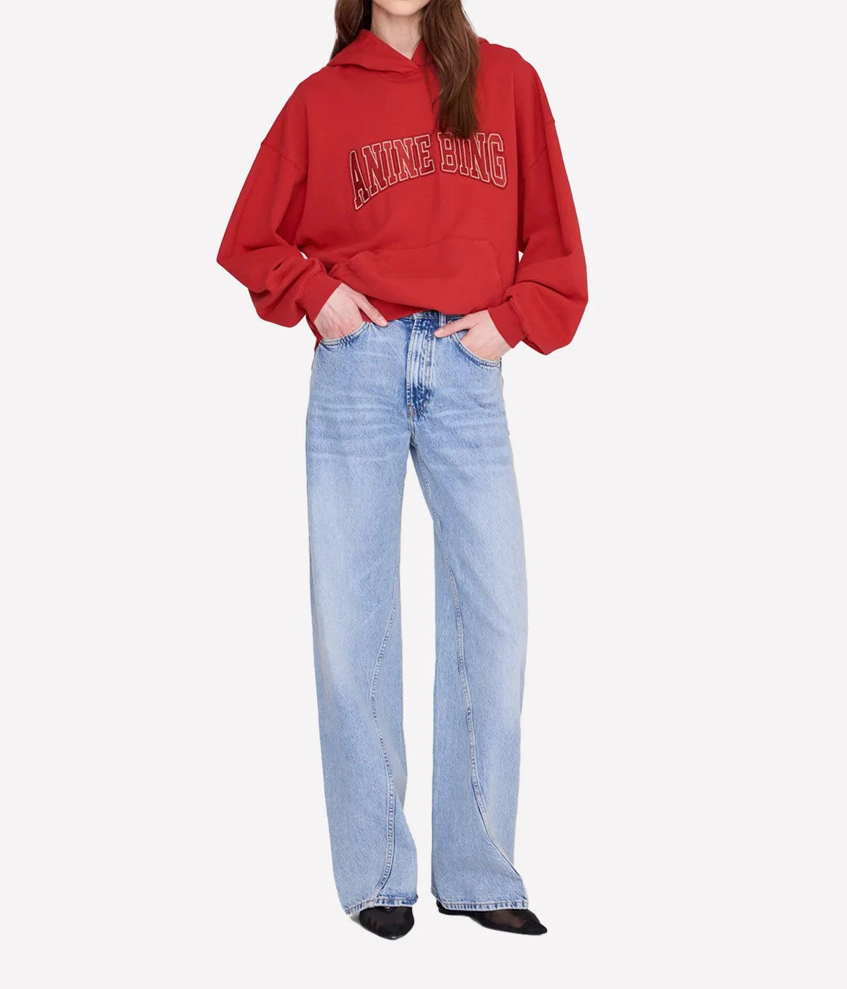 A model wearing a red cropped hoodie with 'ANNE KUNG' printed in arched varsity letters on the chest, paired with light-wash wide-leg jeans and black pointed flats, against a plain light gray background.