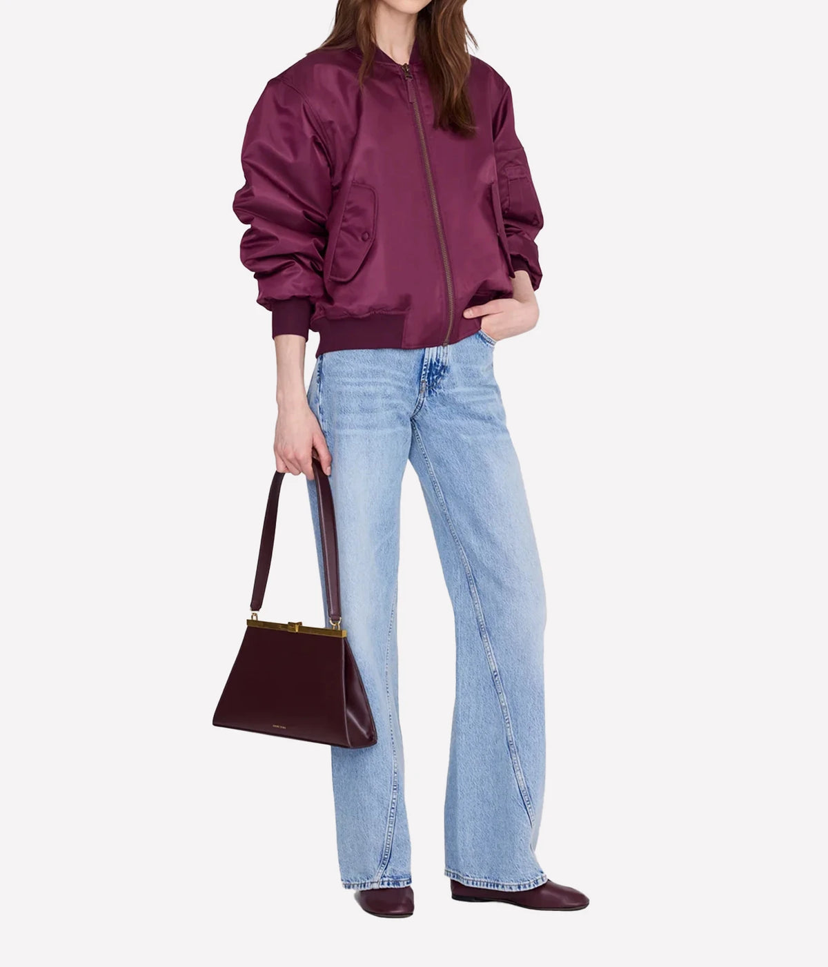 A model wearing a deep burgundy satin bomber jacket, light-wash wide-leg jeans, and matching burgundy flats, holding a structured burgundy handbag against a plain light gray background.