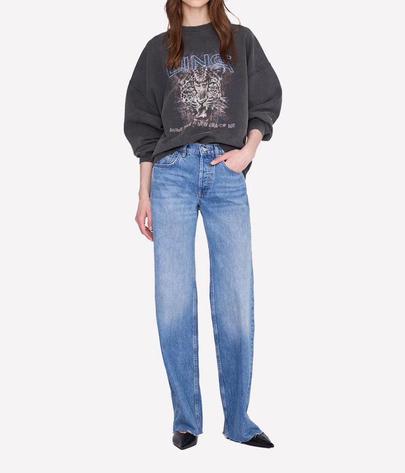 A model wearing a dark gray graphic sweatshirt and light blue wide-leg jeans, standing against a light gray studio background.