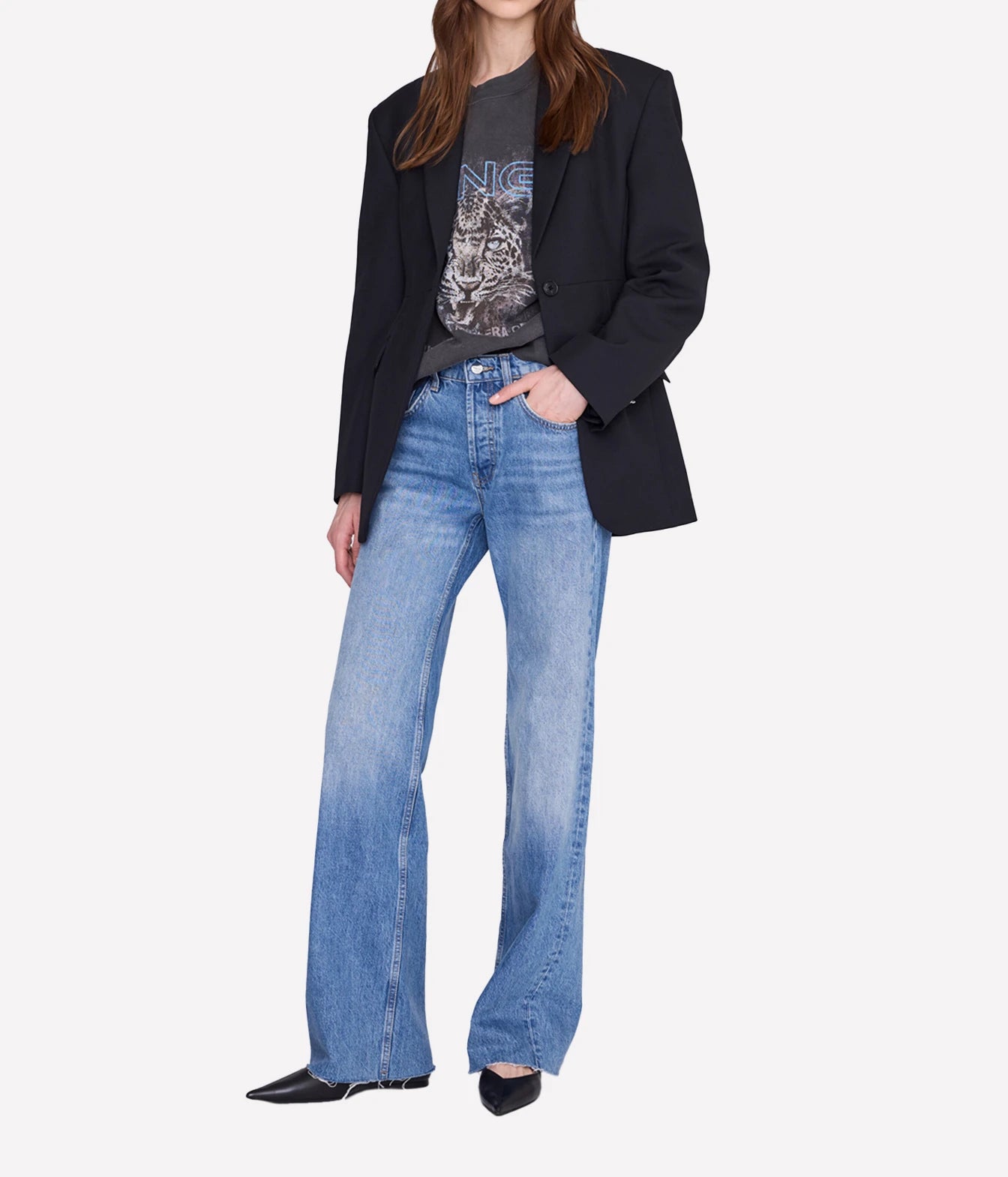 A model wearing a black blazer, a dark graphic t-shirt, and light-wash blue wide-leg jeans, standing against a plain light gray background.