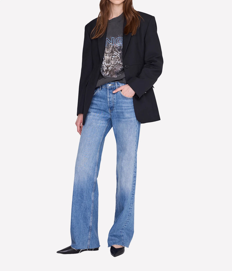 A model wearing a black blazer, a dark graphic t-shirt, and light-wash blue wide-leg jeans, standing against a plain light gray background.