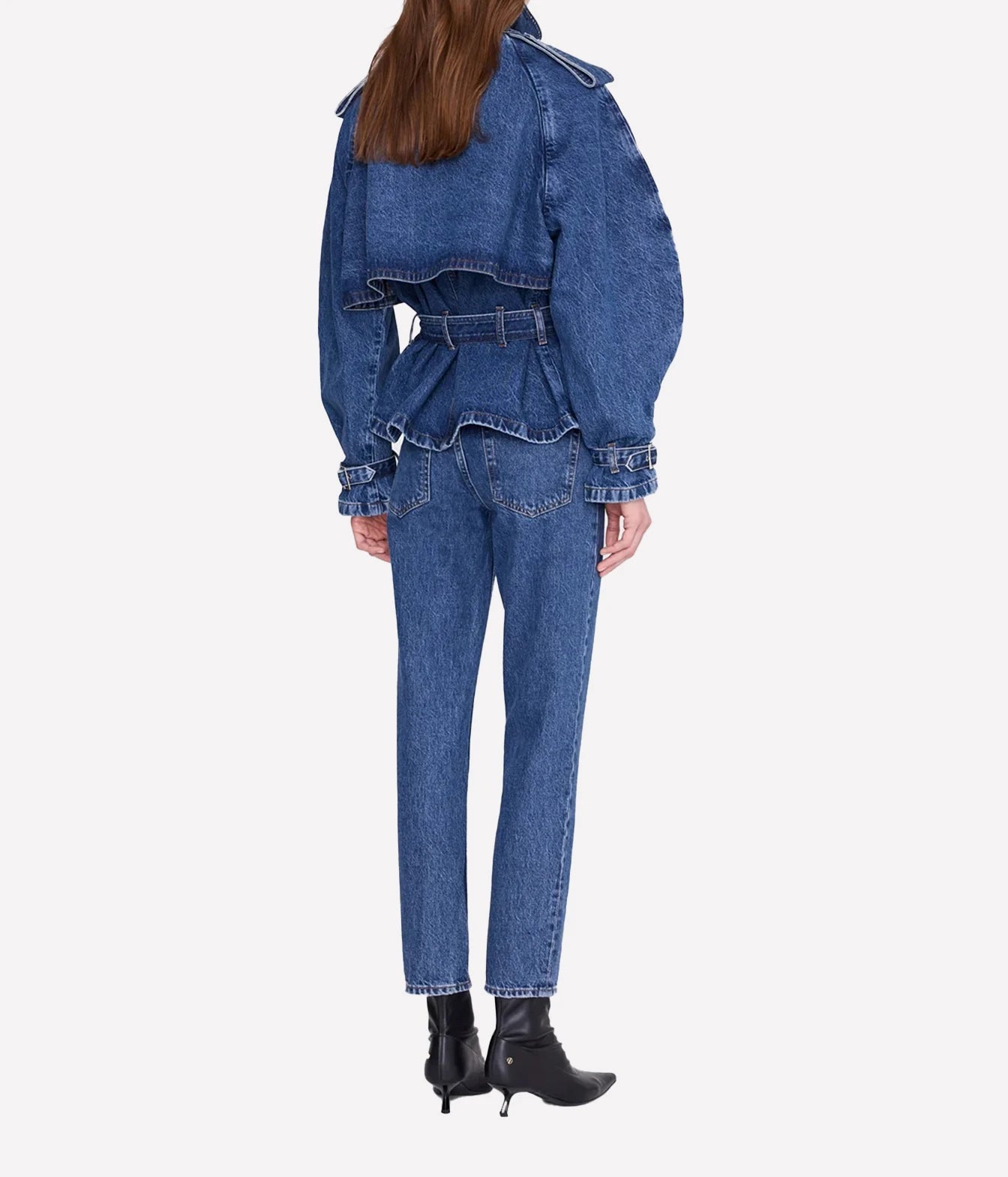 A model shown from the back wearing a blue denim set consisting of a cropped belted jacket with dramatic puffed sleeves and matching straight-leg jeans, paired with black ankle boots, on a light gray background.