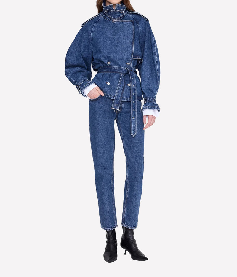 A model wearing a blue denim one-piece jumpsuit with a belted waist, high collar, and long sleeves, paired with black heeled ankle boots against a light gray background.