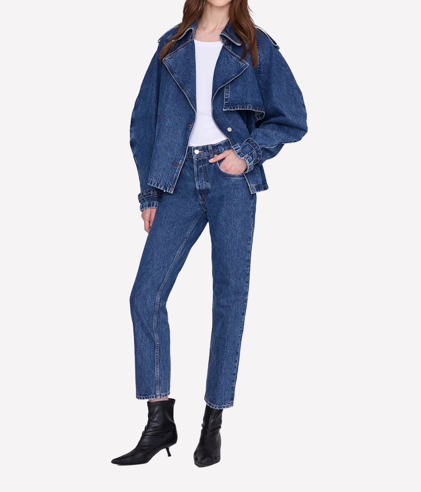 A model wearing a blue denim jacket and matching blue jeans with a white top and black ankle boots, shown against a plain light gray studio background.
