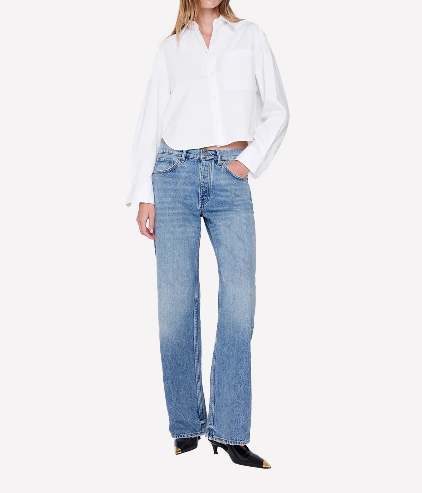 A model wearing light-wash blue straight-leg jeans, a white button-down shirt, and black pointed-toe heels with gold tips, photographed against a clean off-white studio background.
