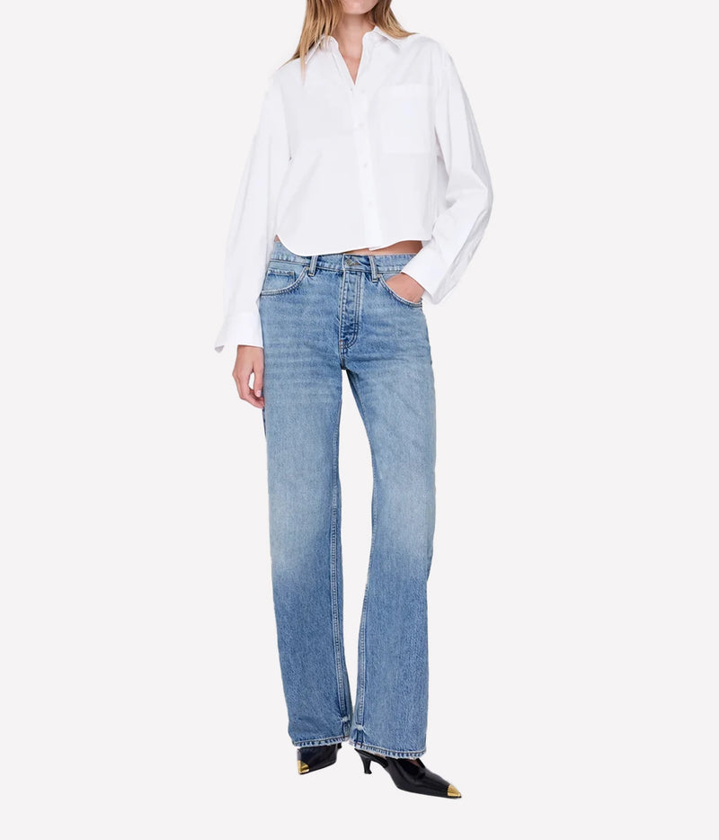 A model wearing light-wash blue straight-leg jeans, a white button-down shirt, and black pointed-toe heels with gold tips, photographed against a clean off-white studio background.