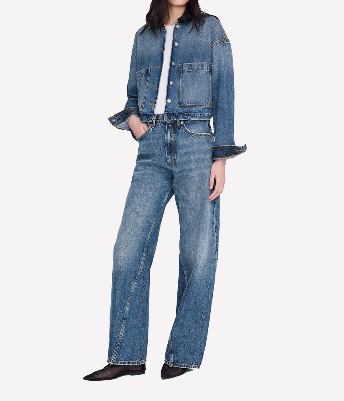 A model wearing a light blue cropped denim jacket with rolled cuffs, matching wide-leg blue jeans, a white top, and black pointed shoes, photographed against a light gray studio background.