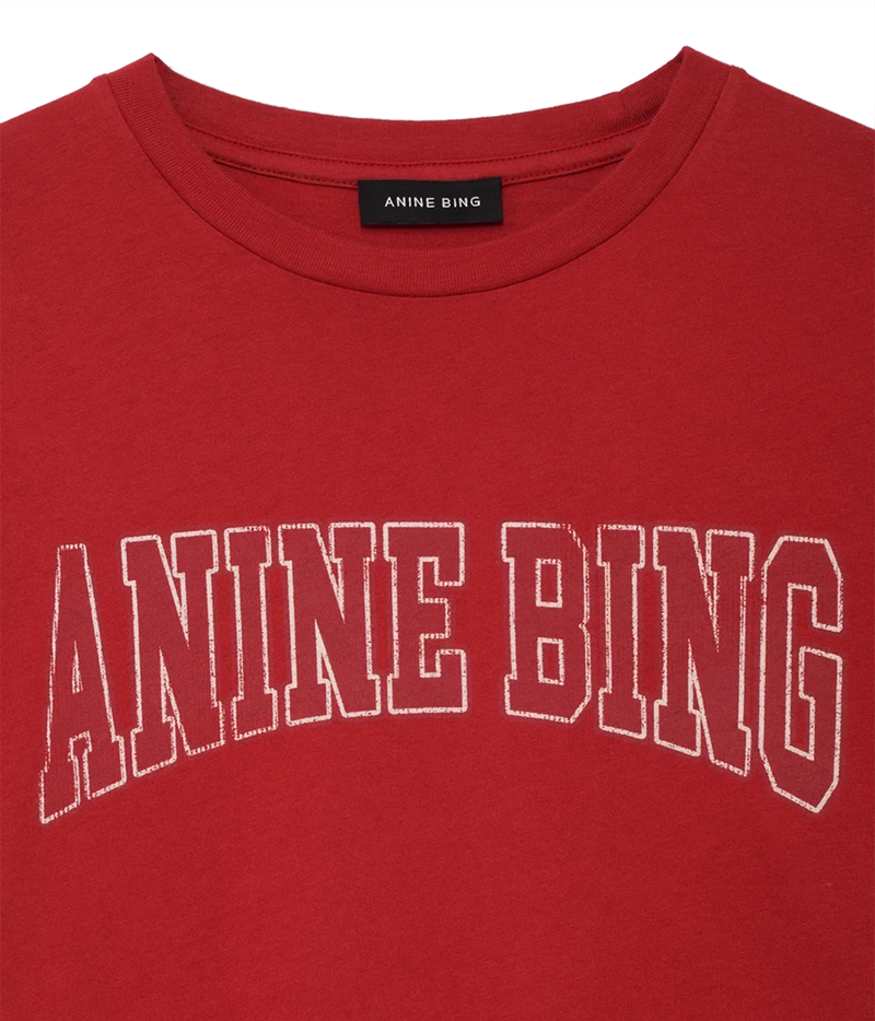 A close-up of a red crewneck t-shirt featuring the brand name 'ANINE BING' printed in white outline block letters across the chest. The black neck label also displays the brand name.