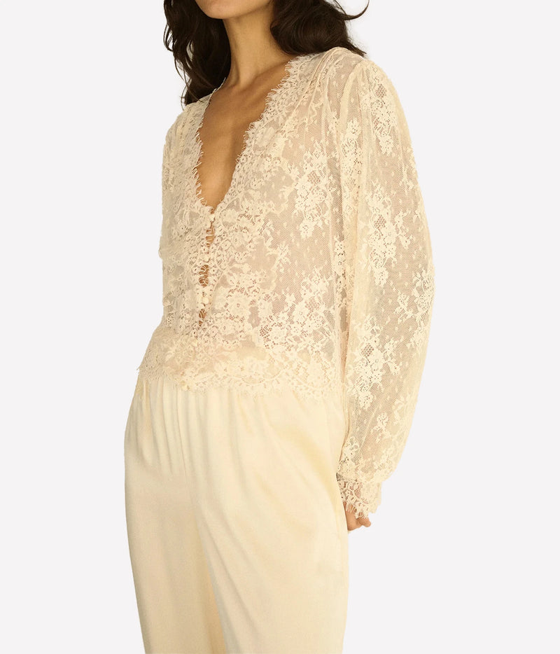 Three-quarter view of a model wearing the DÔEN Amaline Top In Candlelight with cream satin trousers, showing the sheer lace body and cuffed sleeves