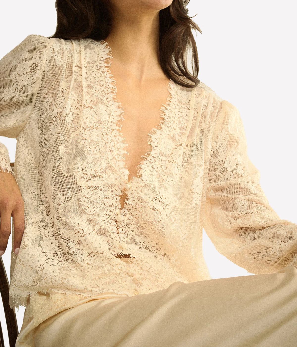 Close-up of a seated model wearing the DÔEN Amaline Top In Candlelight with cream satin trousers, showing the deep V-neck and lace sleeves