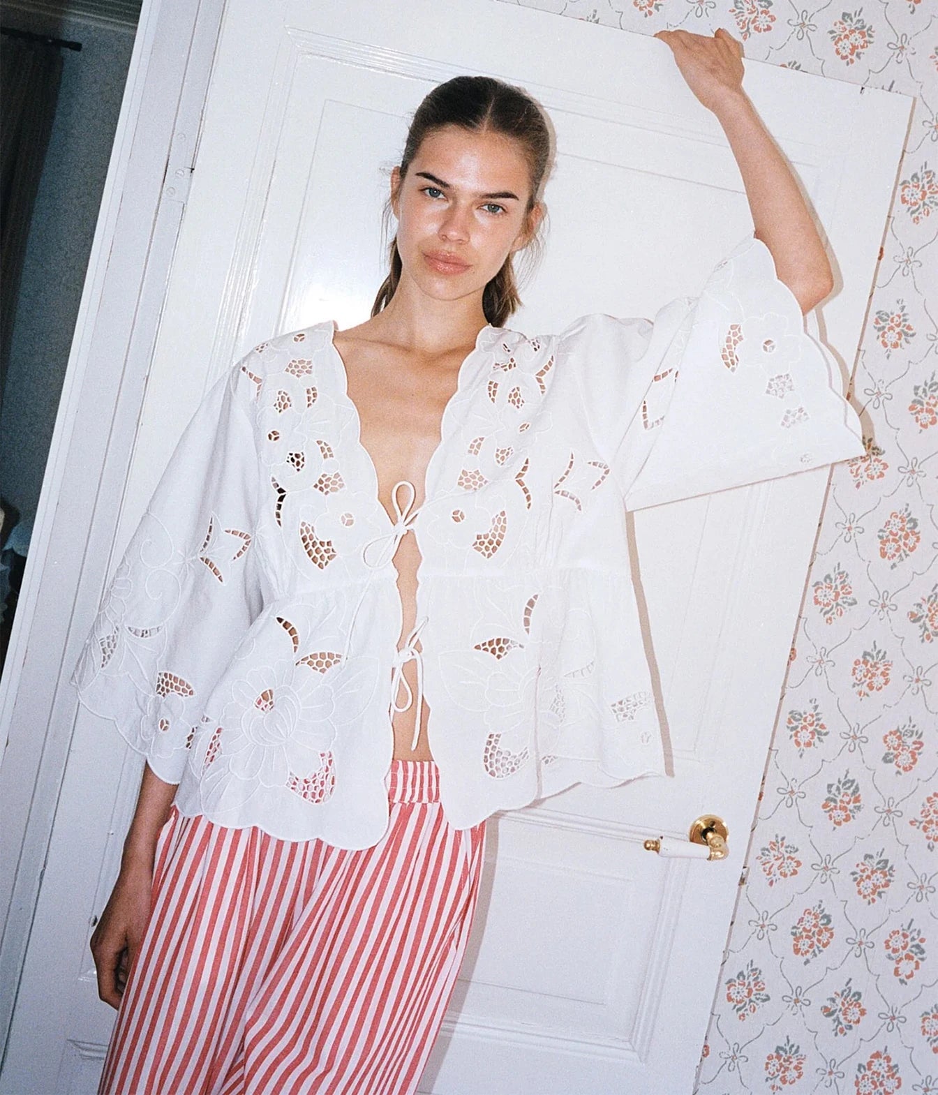 A woman wearing a white eyelet lace cropped cardigan with wide bell sleeves and red-and-white vertically striped pants, posing in front of a white door with patterned wallpaper.