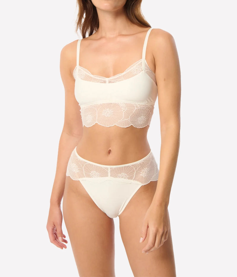 A model wearing a two-piece lingerie set in a warm off-white/cream color, featuring a bralette with floral lace trim and matching high-waisted briefs, Anemone Cropped Cami In Ivory