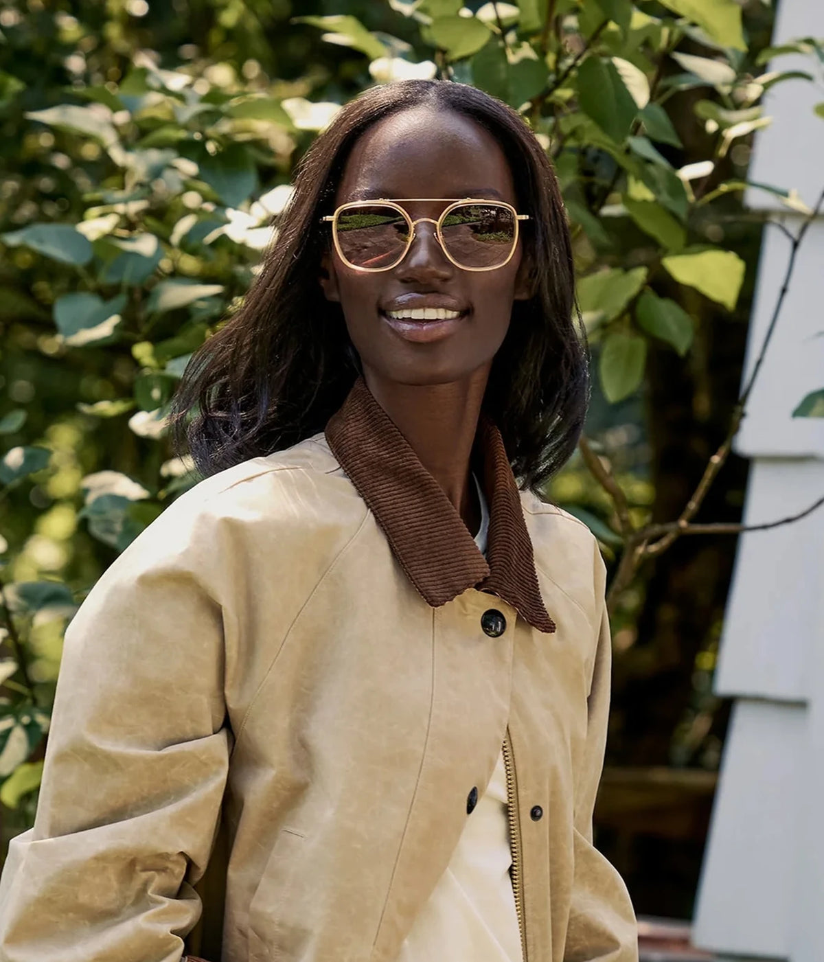 Model smiling outdoors against greenery, wearing the KREWE Austin Sunglasses in 18K Titanium Champagne with a beige jacket