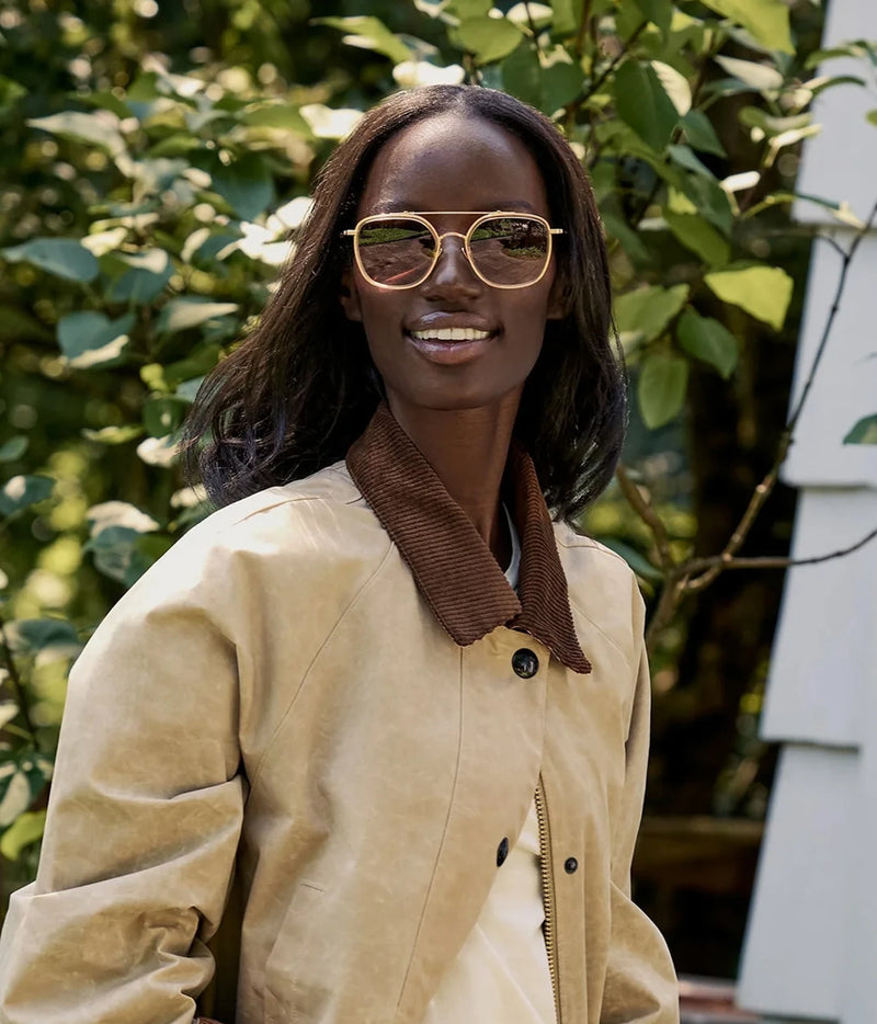 Model smiling outdoors against greenery, wearing the KREWE Austin Sunglasses in 18K Titanium Champagne with a beige jacket