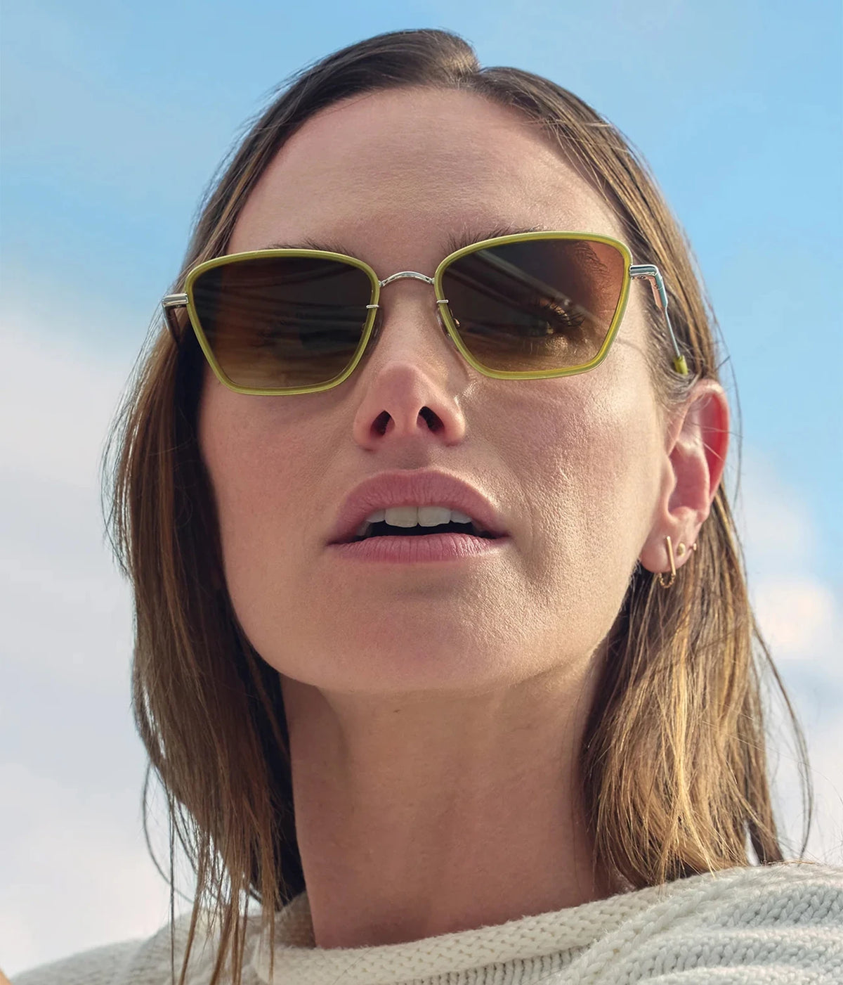 Model outdoors against a blue sky, wearing the KREWE Bella Sunglasses in Silver Chartreuse with a cream top