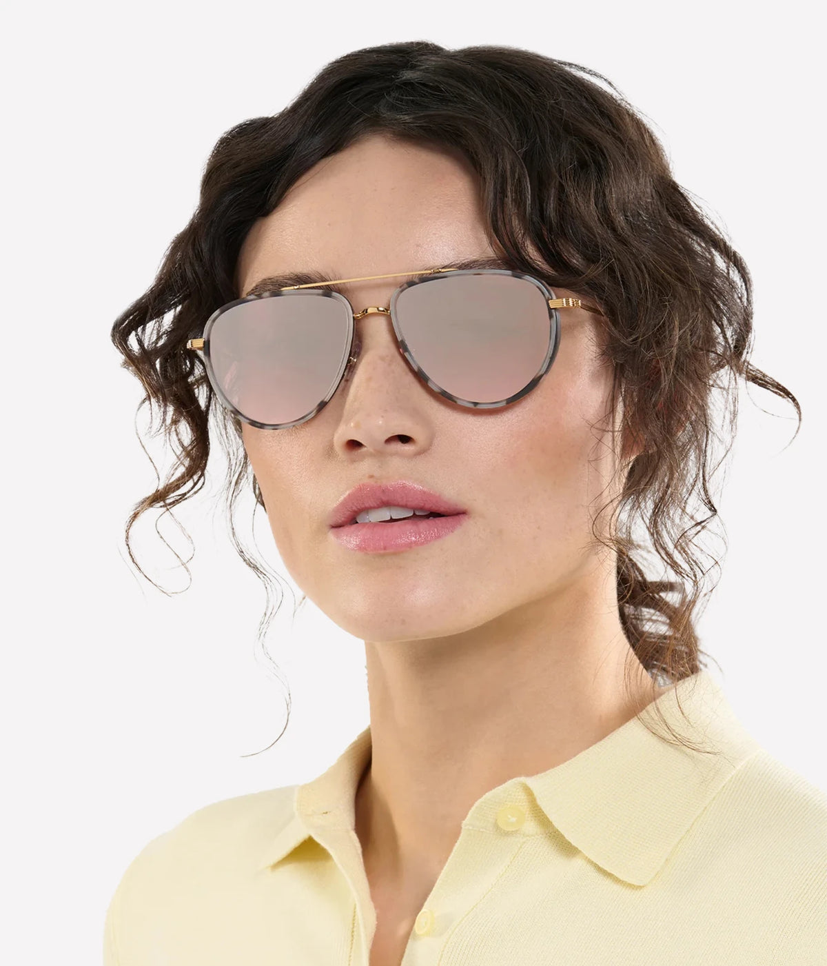 Model with curly dark hair wearing the KREWE Coleman Sunglasses in Matte Black Tortoise with a pale yellow collared top