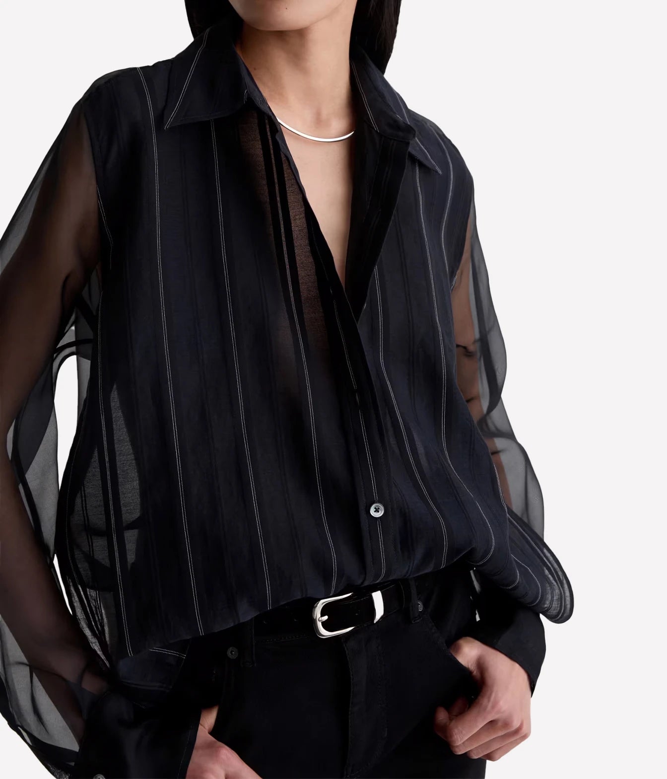 A close-up of a model wearing a sheer black long-sleeve blouse with vertical pinstripes and a collar, paired with black trousers and a silver belt.