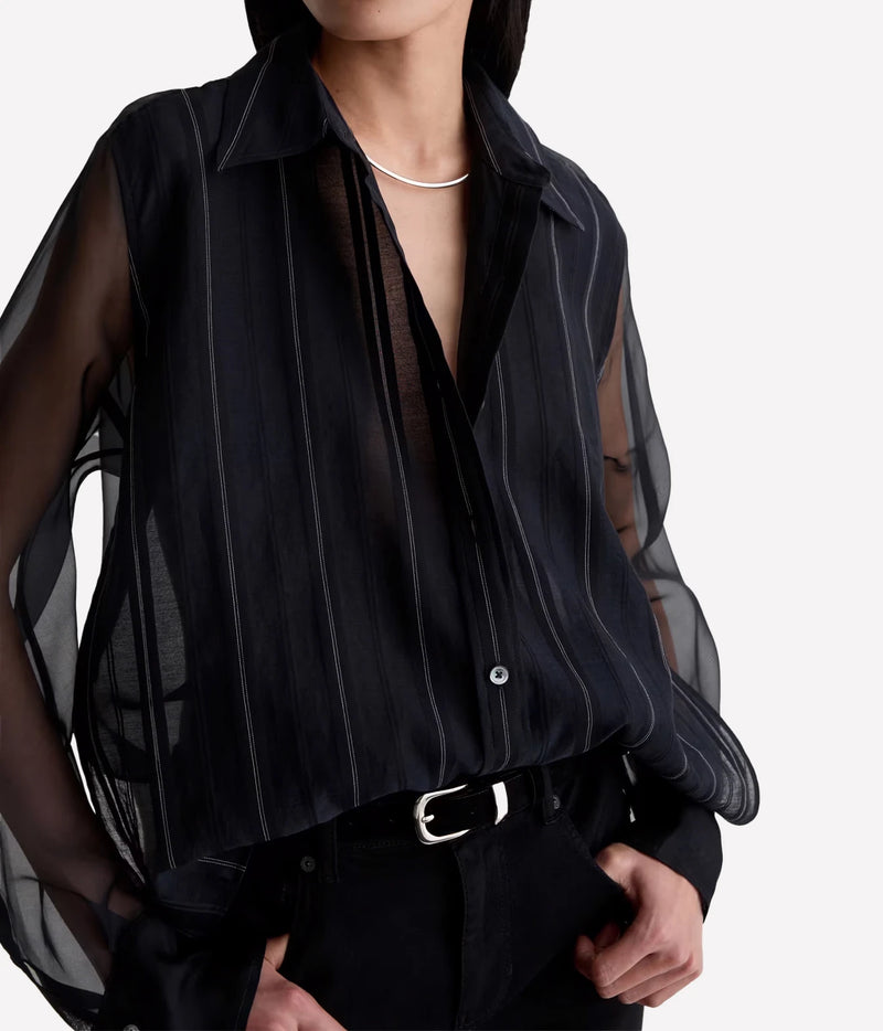 A close-up of a model wearing a sheer black long-sleeve blouse with vertical pinstripes and a collar, paired with black trousers and a silver belt.