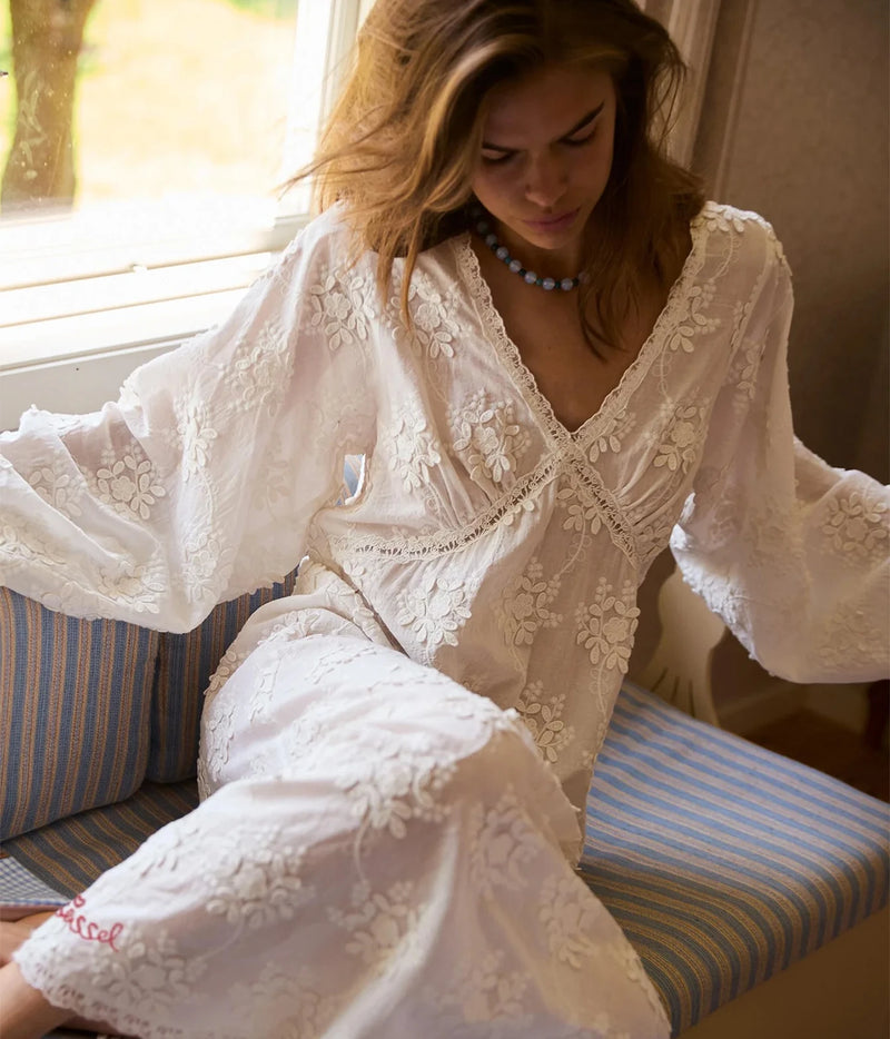 A woman wearing a white embroidered lace robe sits on a blue and white striped cushioned bench near a window.