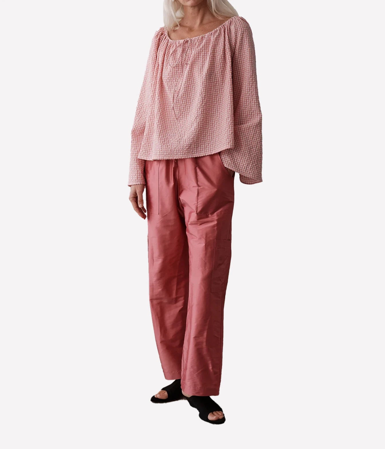 A model wearing a dusty pink two-piece set consisting of a loose, off-the-shoulder gingham long-sleeve top and matching wide-leg trousers, paired with black slide sandals, against a plain light gray background.