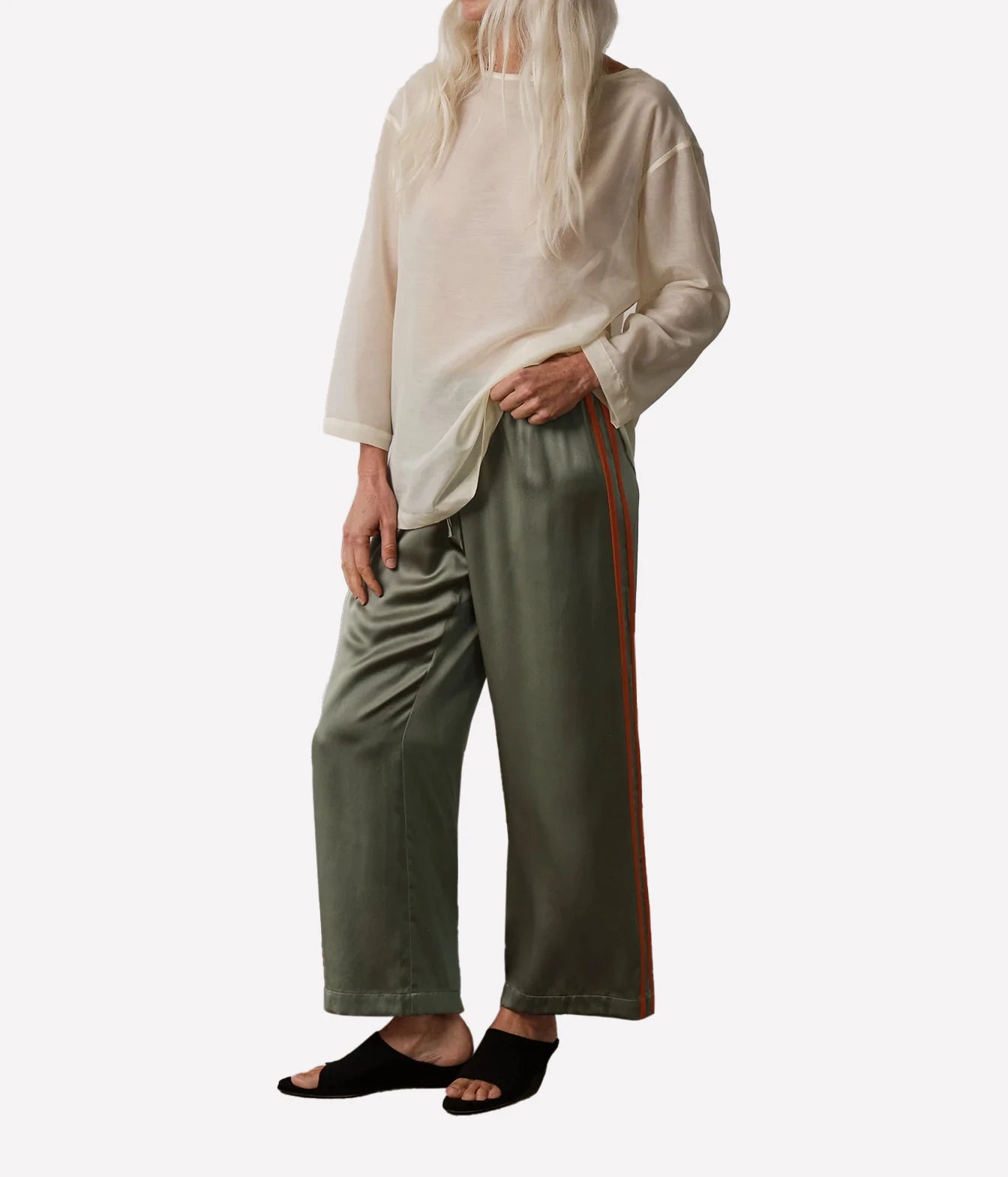A model wearing wide-leg sage green pants with a rust-orange stripe down the side, paired with a loose cream-colored long-sleeve top and black slide sandals, set against a plain light gray background.