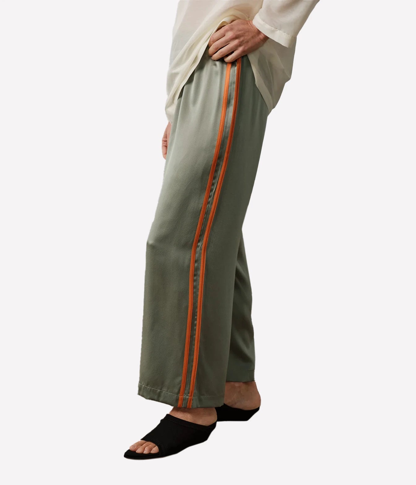 A person wearing wide-leg sage green pants with a double orange stripe down the side, paired with a cream top and black slide sandals, shown against a plain light gray background.