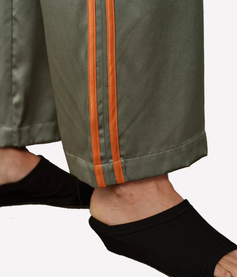 Close-up of olive green pants with two vertical orange stripes down the side, worn with black slip-on shoes against a light gray background.