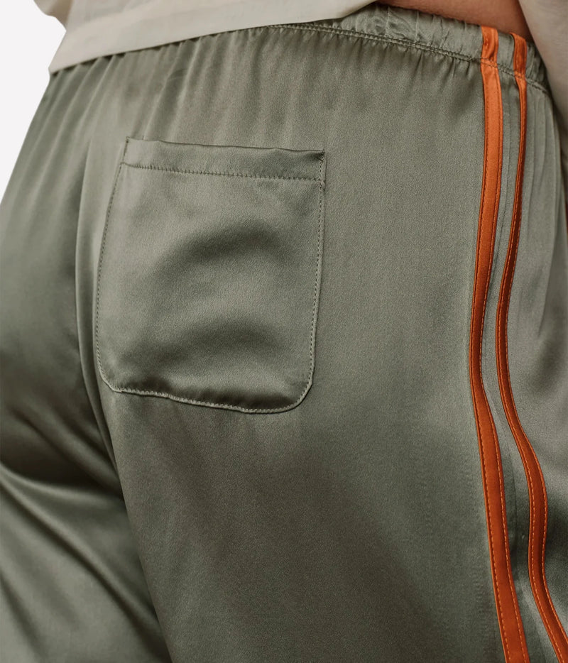 Close-up of olive green satin-like track pants featuring a side pocket and double orange piping along the side seam.