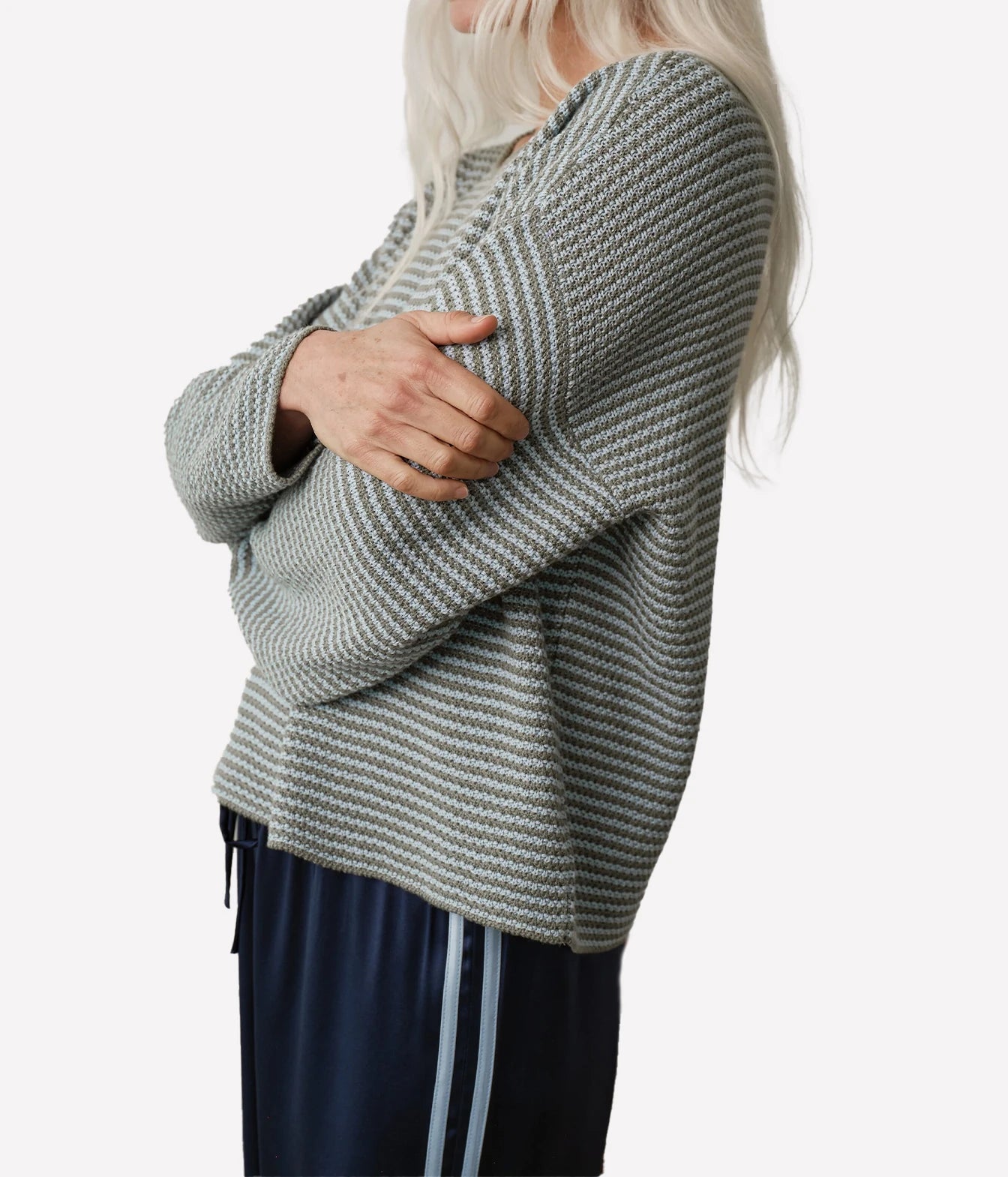 A woman wearing a textured knit sweater with horizontal gray and white stripes, paired with navy blue track pants with white side stripes, shown from a side profile against a light gray background.