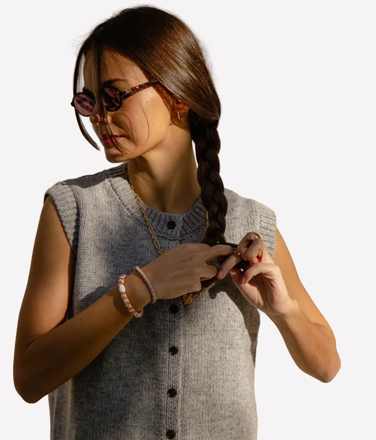A woman with long brown hair wearing a grey sleeveless button-front knit top and tortoiseshell sunglasses, adjusting her hair braid